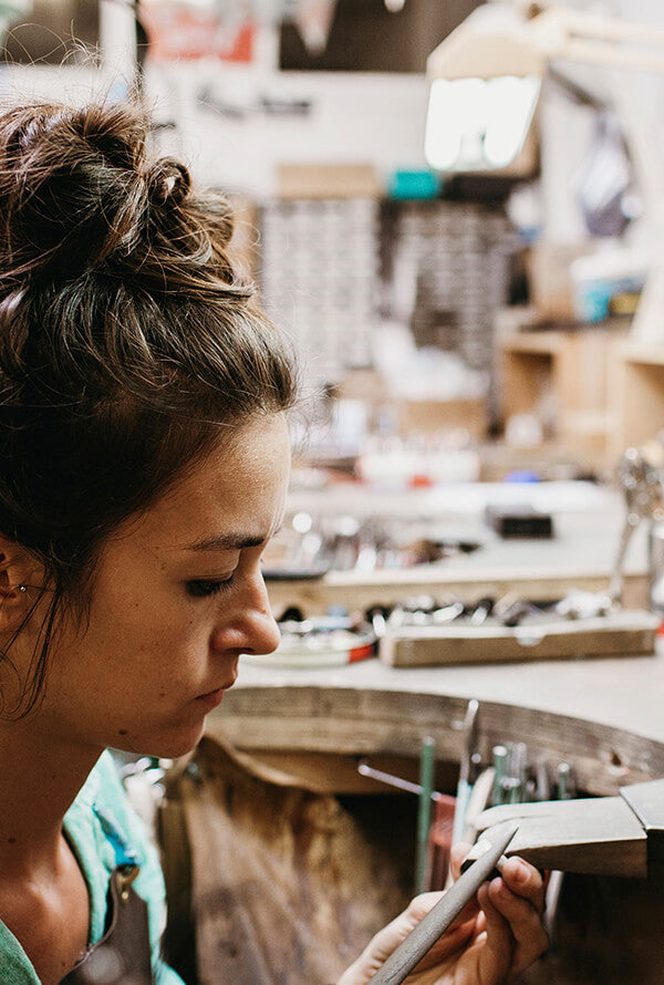 A picture of a female jeweller working at her bench. In the background we can see lots of tools. She is concentrating on her work.